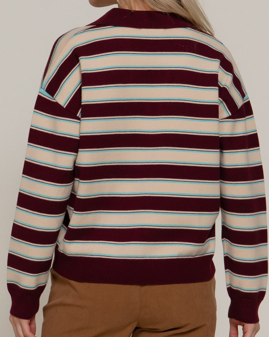 A woman shown from the back wearing a long-sleeve knit sweater with horizontal stripes in burgundy, cream, and light blue, paired with brown pants, against a plain beige background.