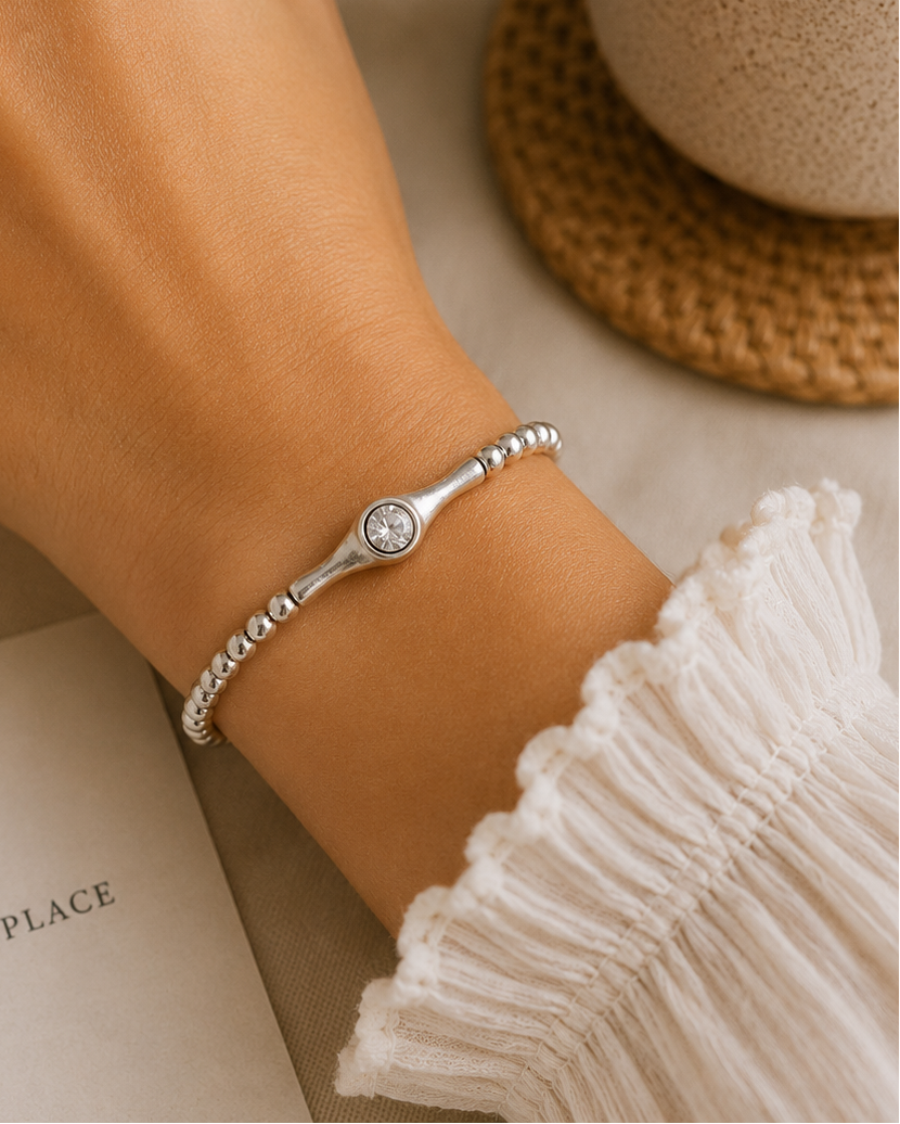 Hand wearing a silver bracelet with a small round gemstone, next to a cup and book on a textured surface.