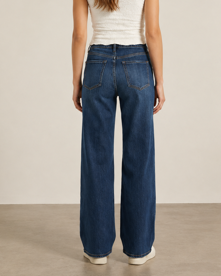 A woman viewed from the back wearing high-waisted, wide-leg blue jeans, a white ribbed tank top, and white sneakers, standing against a neutral beige studio background.
