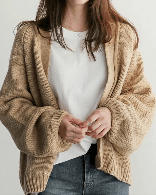 Person wearing a beige cardigan over a white shirt and jeans on a plain background