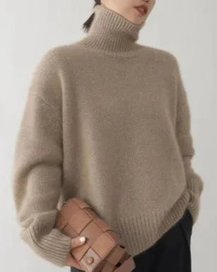 Person wearing a beige turtleneck sweater holding a brown leather bag against a plain background