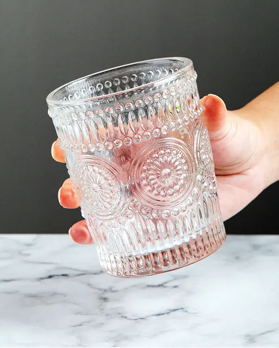 Hand holding a decorative glass tumbler on a marble surface with a dark background