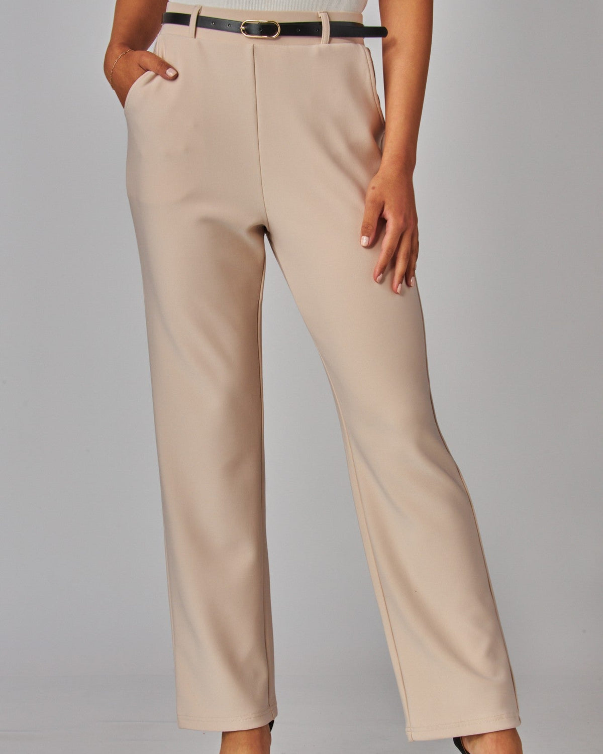 A model wearing high-waisted, straight-leg beige trousers with a black belt, paired with a white top and black heels, against a light gray studio background.