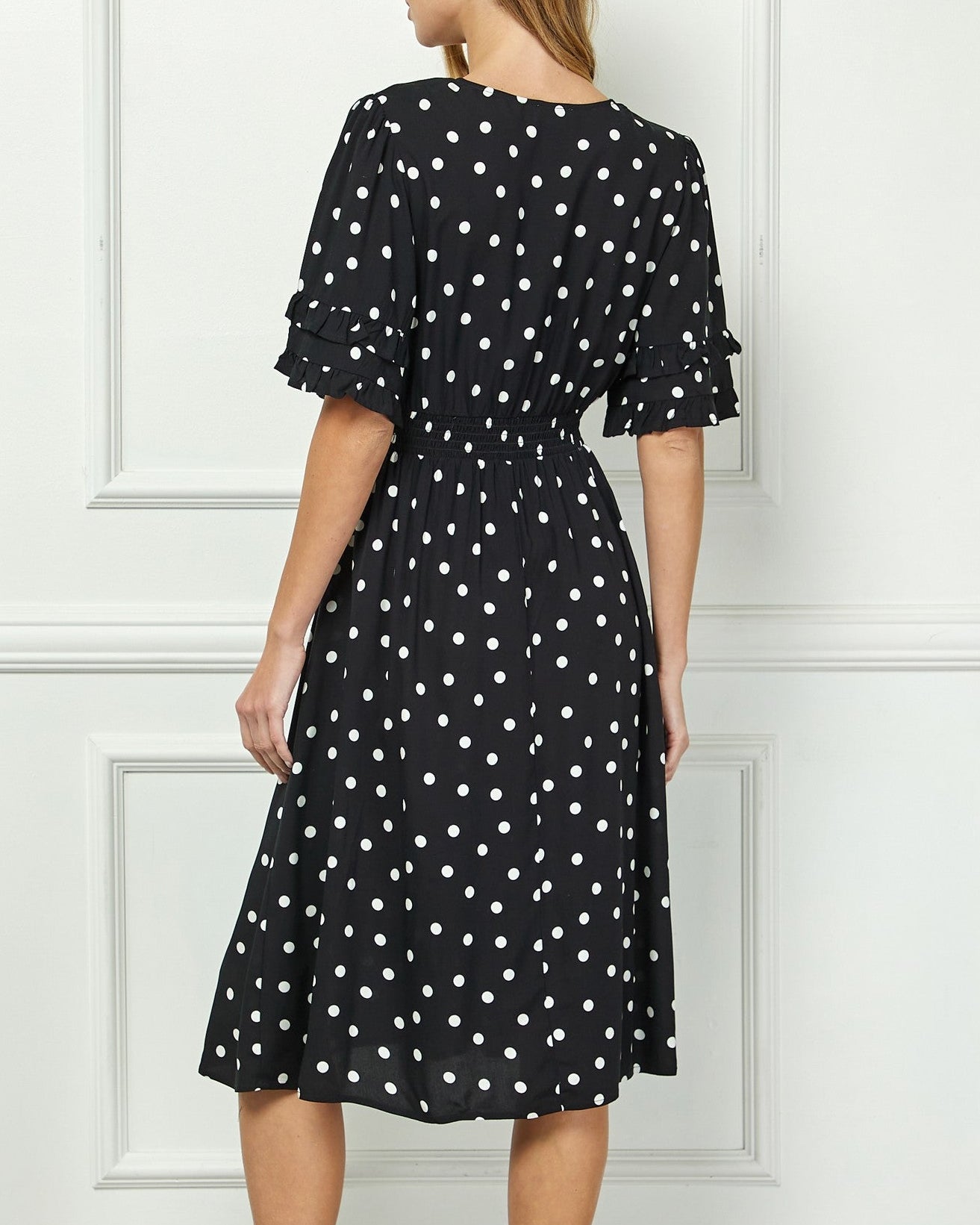 Woman wearing a black dress with white polka dots against a white paneled wall.