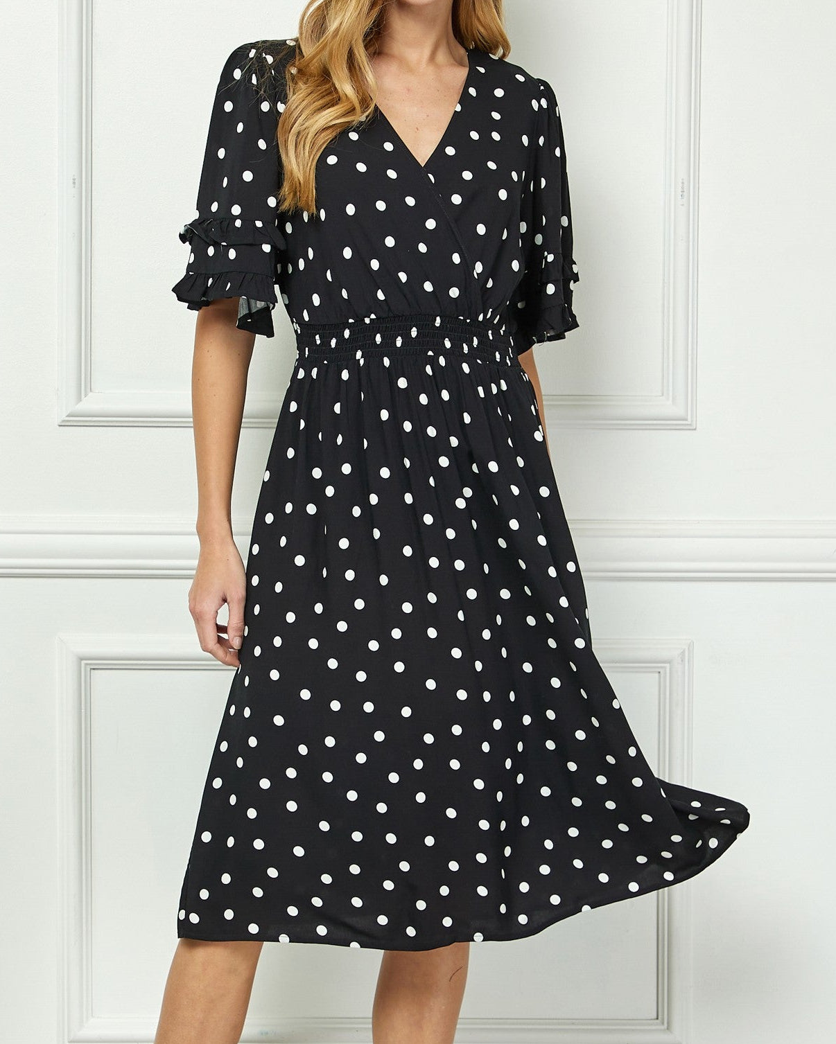 Woman wearing a black dress with white polka dots against a white wall.