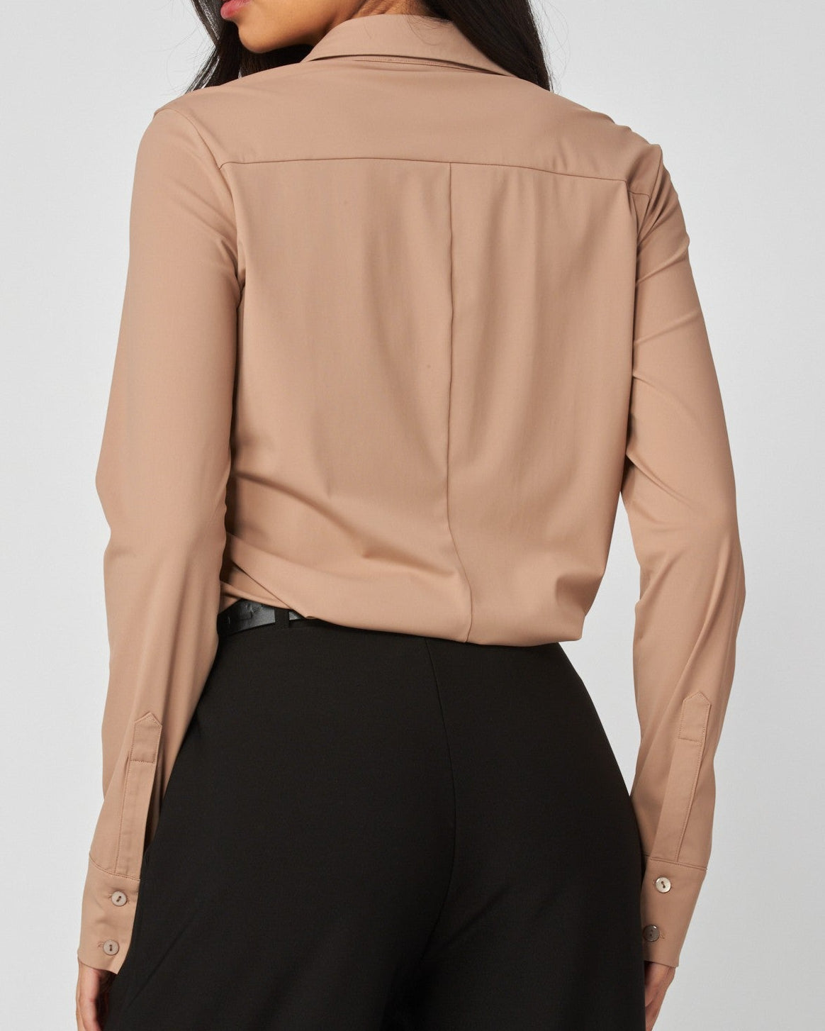 A woman shown from the back wearing a cropped, long-sleeve beige button-up shirt and black trousers, photographed against a light gray studio background.