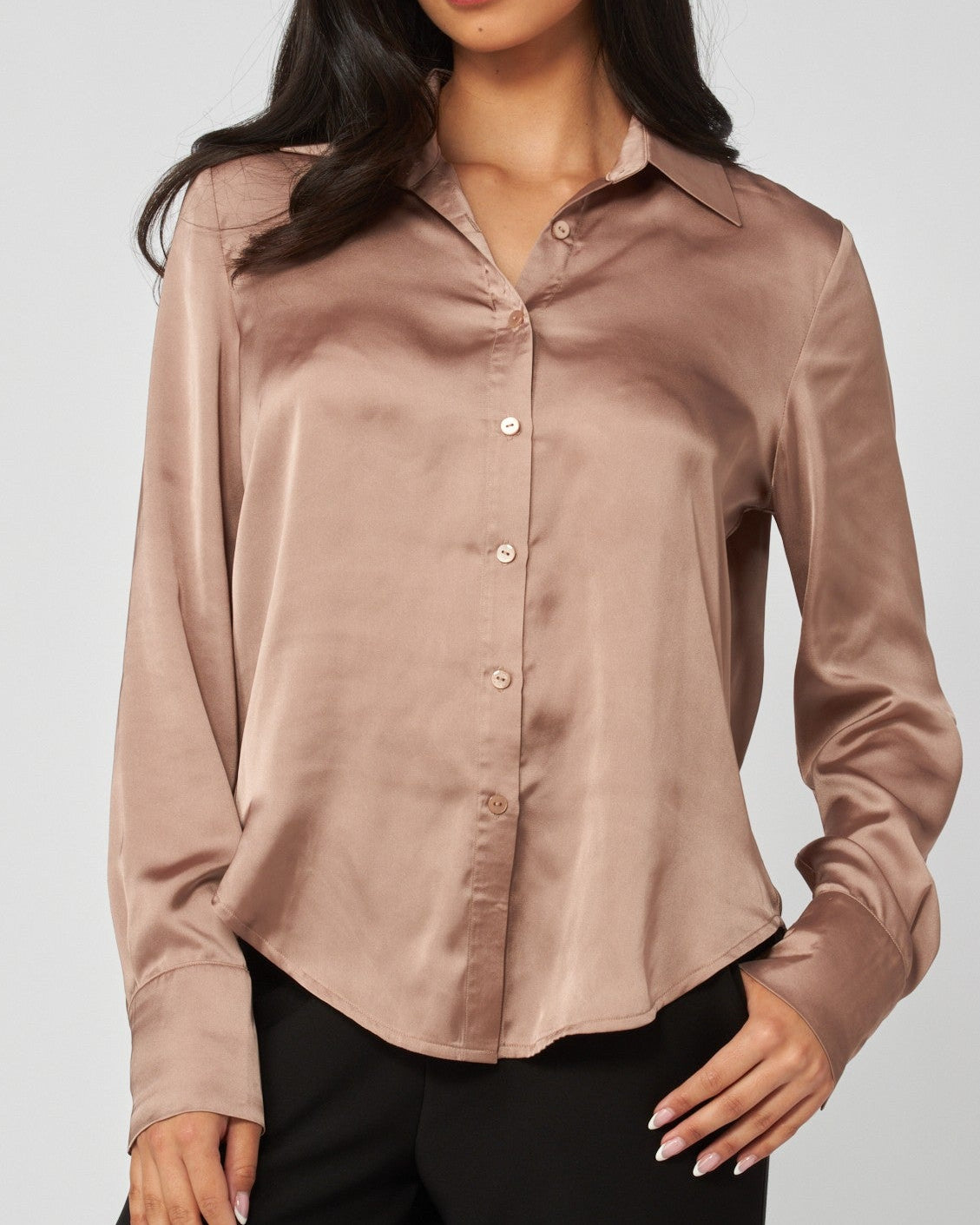 A woman modeling a long-sleeve satin button-up blouse in a dusty rose color, paired with black pants, against a solid light gray background.