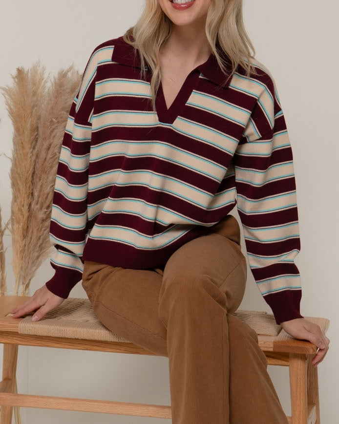 Woman sitting on a wooden bench wearing a striped sweater and brown pants.