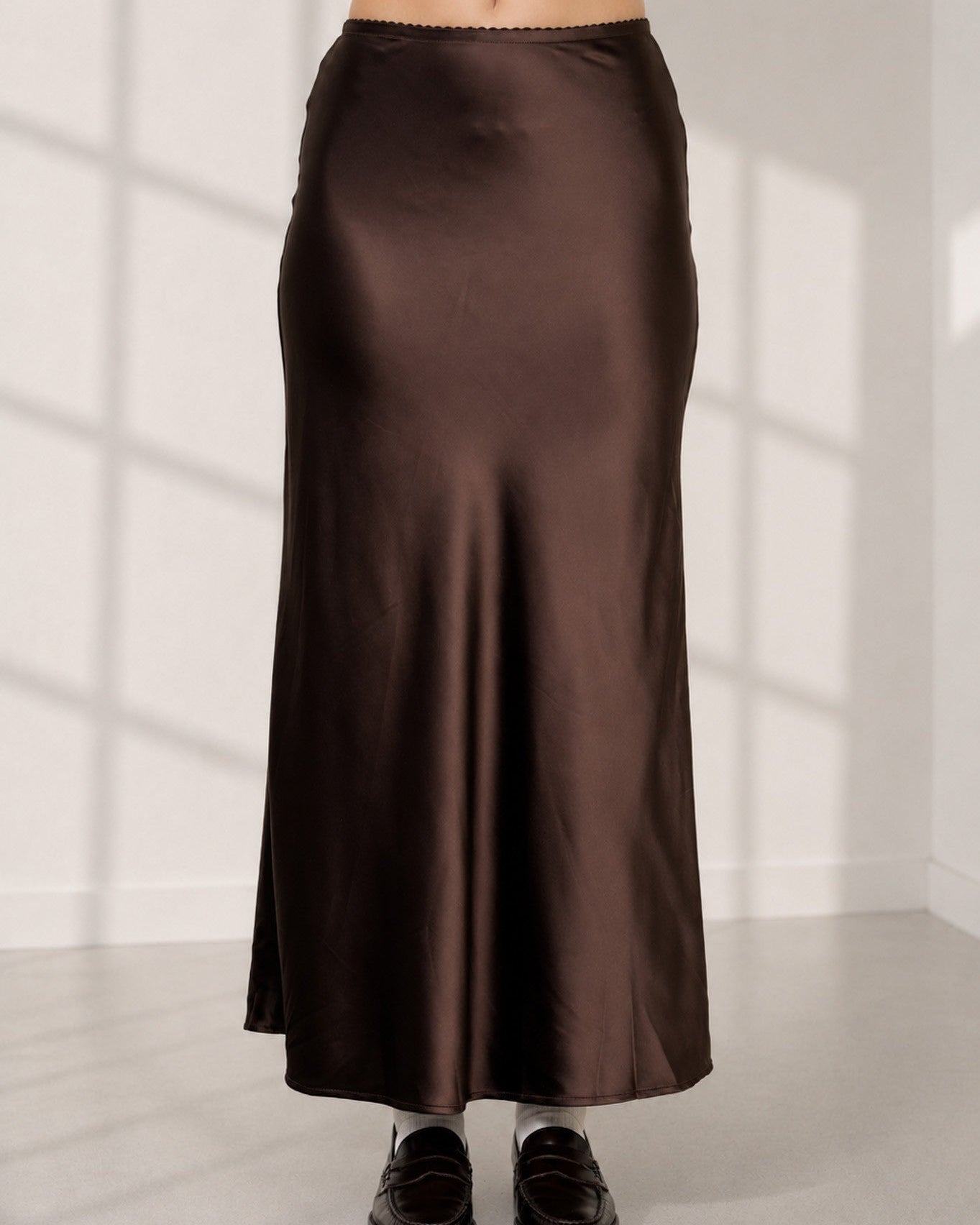 A model is shown from the waist down wearing a dark brown satin midi skirt, a white crop top with black polka dots, and black loafers, standing in a brightly lit room with window pane shadows.