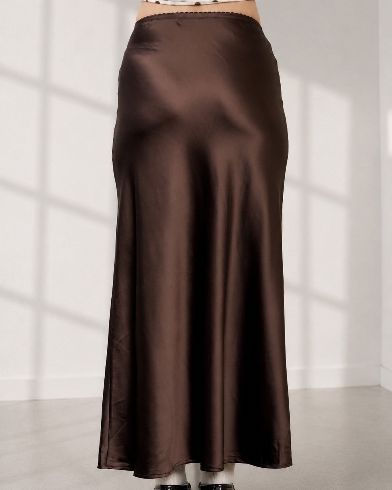 A person wearing a long, satin-like brown skirt with a white polka-dot cropped top and black shoes, standing against a white wall with soft window shadows.