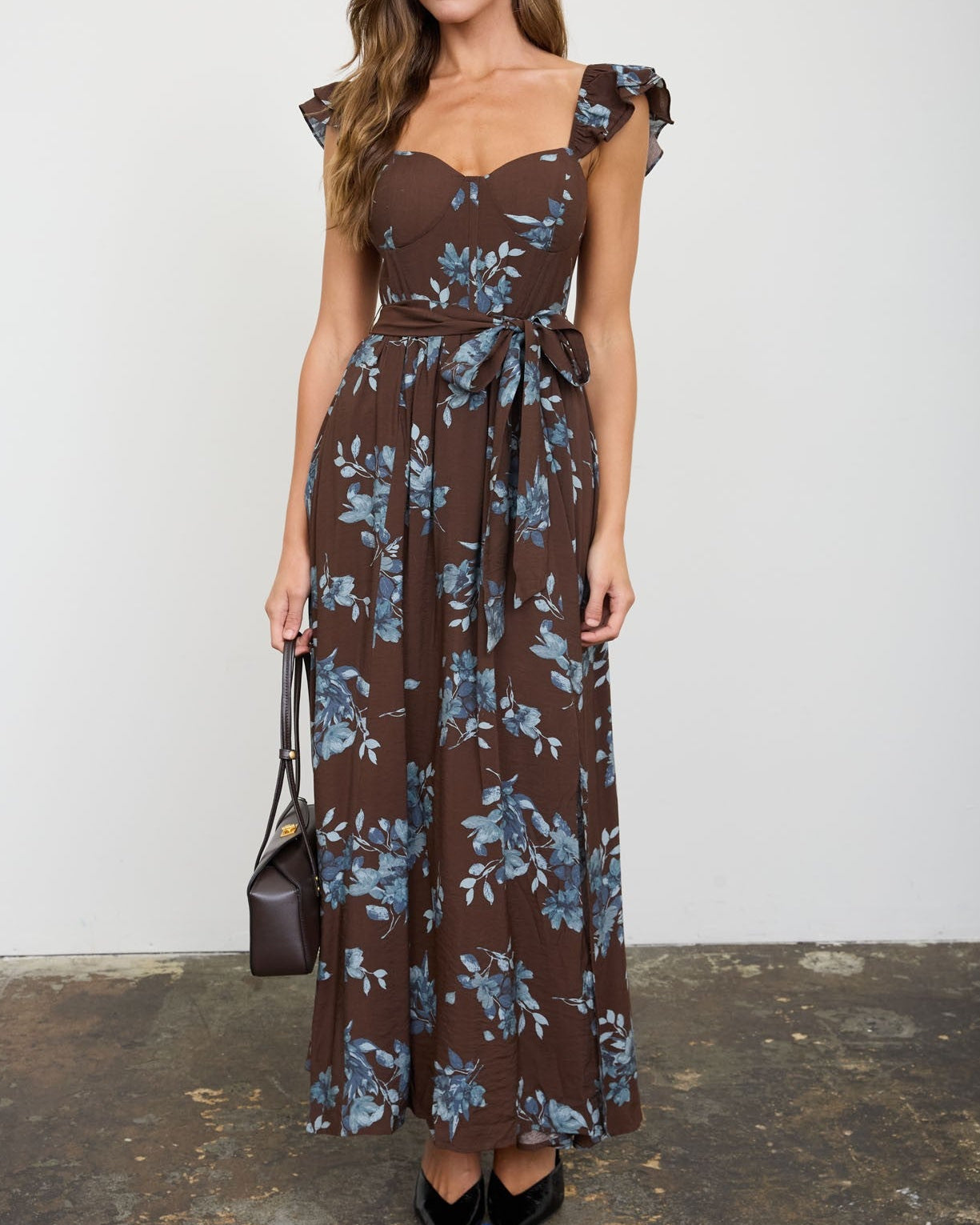 A woman models a brown sleeveless maxi dress with a blue floral print, ruffled cap sleeves, a sweetheart neckline, and a matching waist tie. She is holding a small dark brown handbag and wearing black pointed-toe heels, standing against a white wall with a rustic concrete floor.