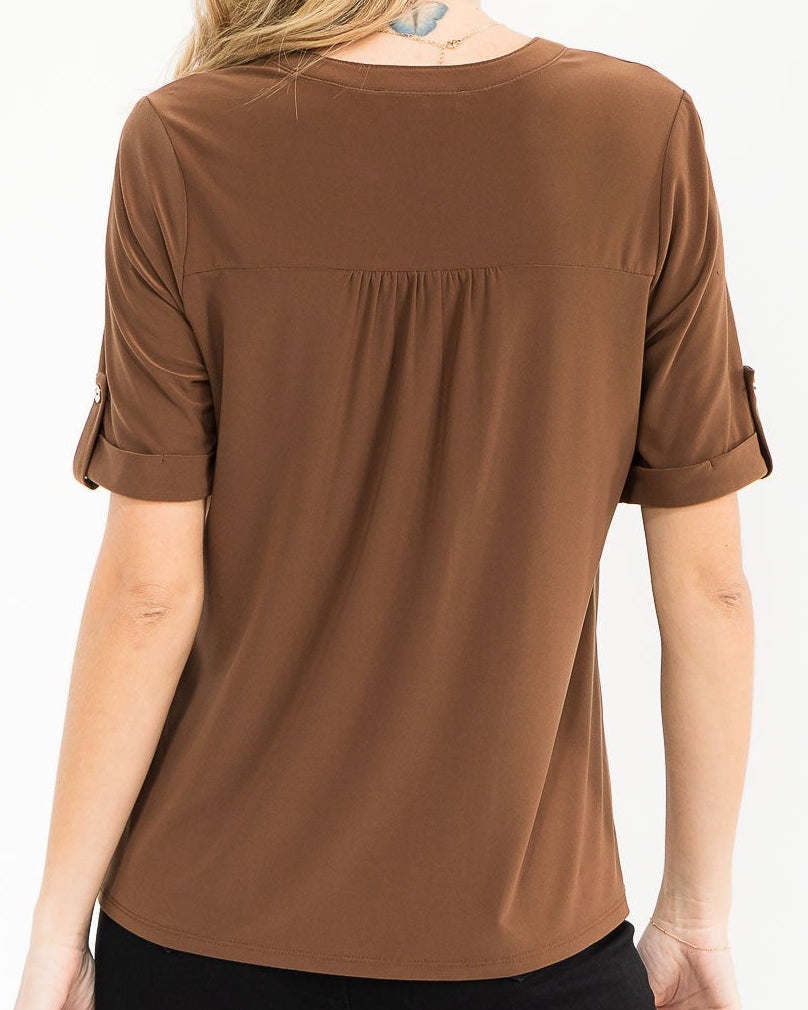 A woman viewed from the back wearing a brown short-sleeve top with rolled cuffs and black pants, against a plain white studio background.