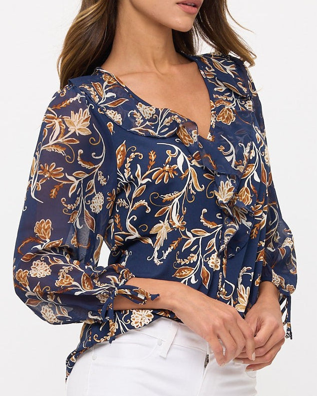 A woman wearing a navy blue long-sleeve blouse with a beige and brown floral pattern, paired with white pants, photographed against a light gray studio background.