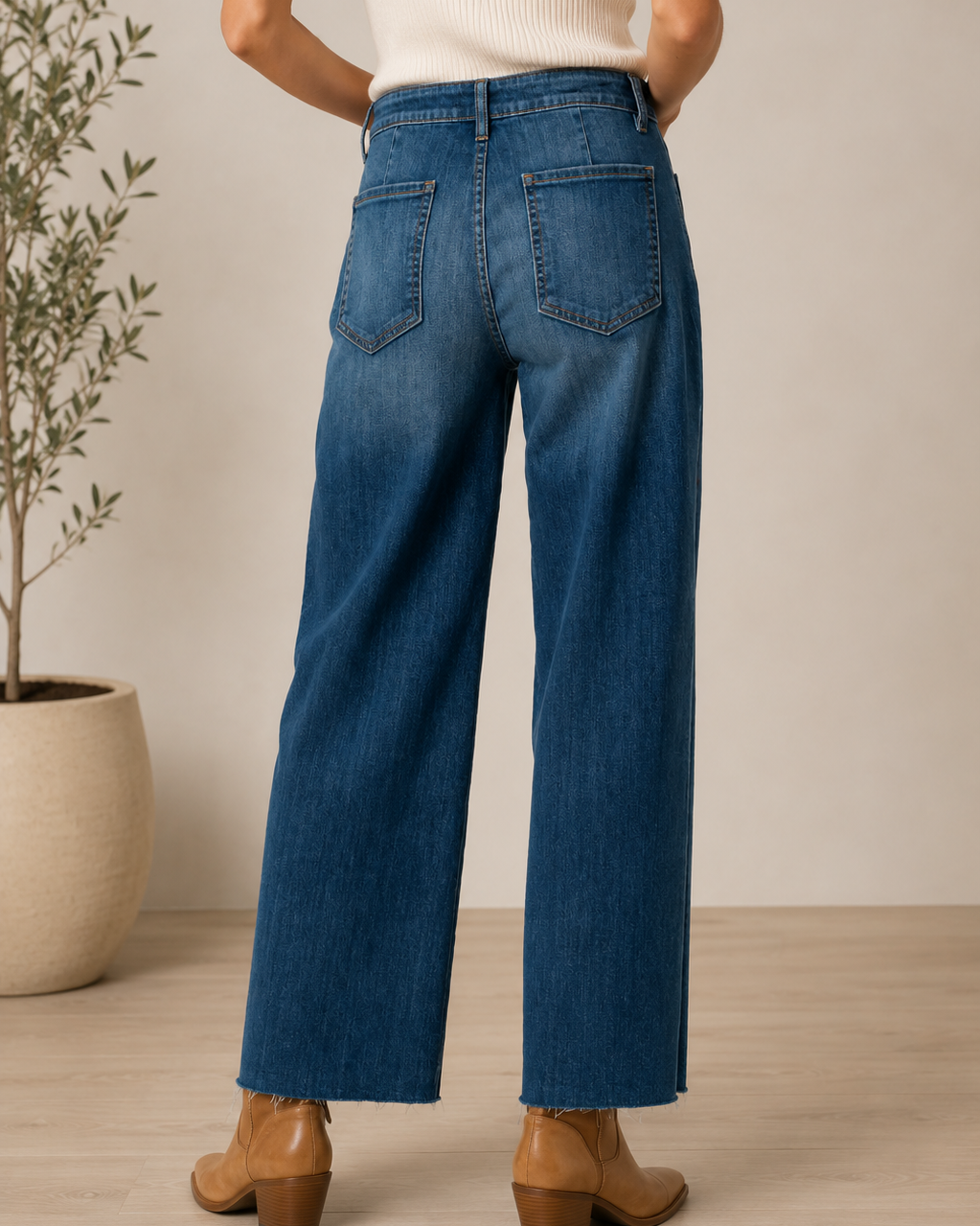 A person shown from the back wearing blue wide-leg jeans, a cream ribbed top, and brown heeled ankle boots in a softly lit indoor setting with a potted plant and light wood floor.
