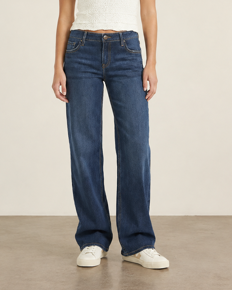 A woman stands in a studio wearing dark blue wide-leg jeans, a white sleeveless knit top, and white sneakers against a plain beige wall and concrete floor.