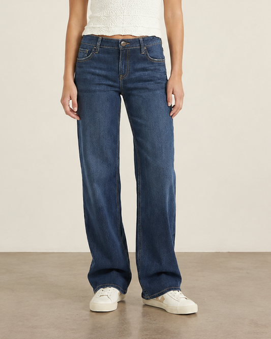 A woman stands in a studio wearing dark blue wide-leg jeans, a white sleeveless knit top, and white sneakers against a plain beige wall and concrete floor.