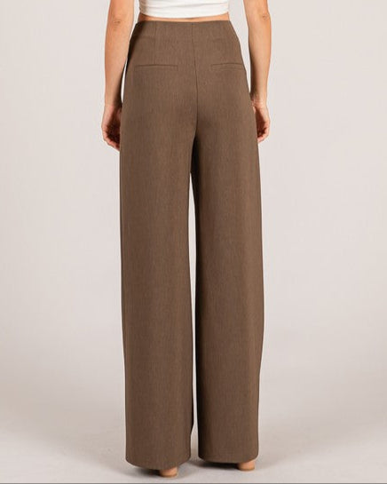Woman wearing brown wide-leg pants and a white top on a plain background