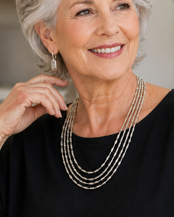 Woman sitting on a couch wearing a black top and multiple necklaces, with a home interior background.