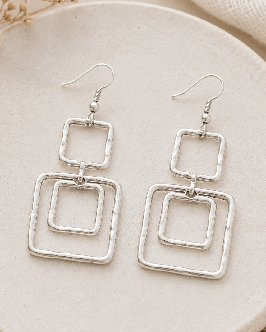 Silver square earrings on a beige plate with dried flowers and fabric.