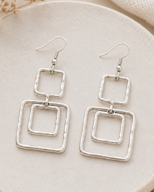 Silver square earrings on a beige plate with dried flowers and fabric.