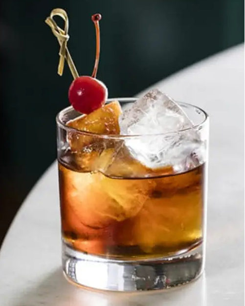 Cocktail with a cherry and lemon peel garnish in a glass on a marble surface.