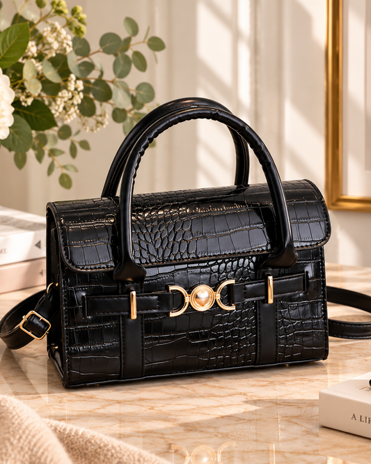 A structured black crocodile-embossed handbag with gold-tone hardware and dual top handles, styled on a marble tabletop with books, flowers, sunglasses, and a small bowl in a warm interior setting.