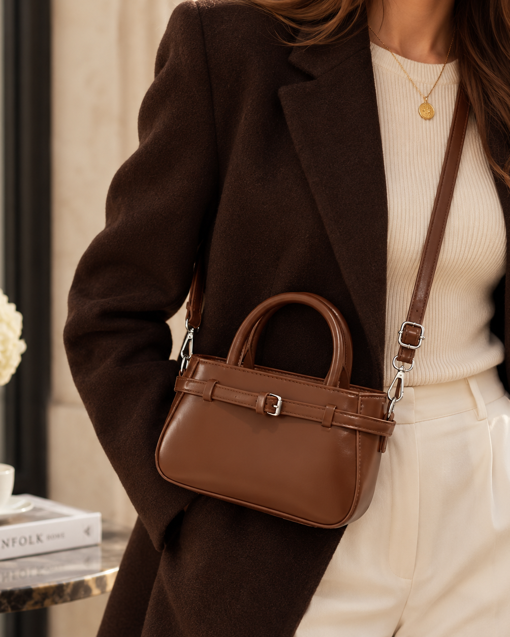 A woman wearing a dark brown blazer and cream ribbed sweater carries a structured brown leather handbag with a top handle and shoulder strap in a softly lit indoor setting.