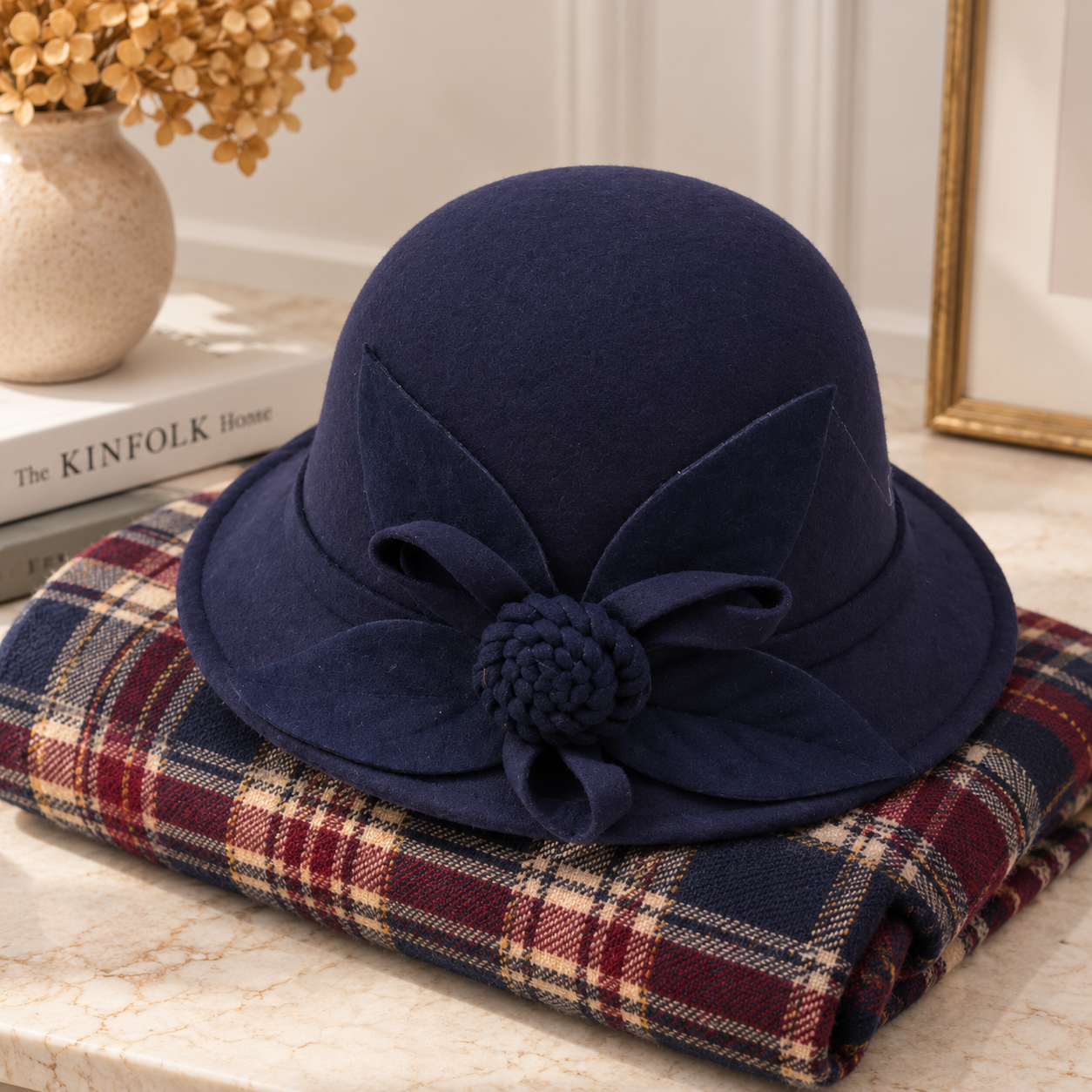 A navy blue felt hat with a large fabric flower on the brim, displayed on top of a folded plaid scarf on a marble surface.