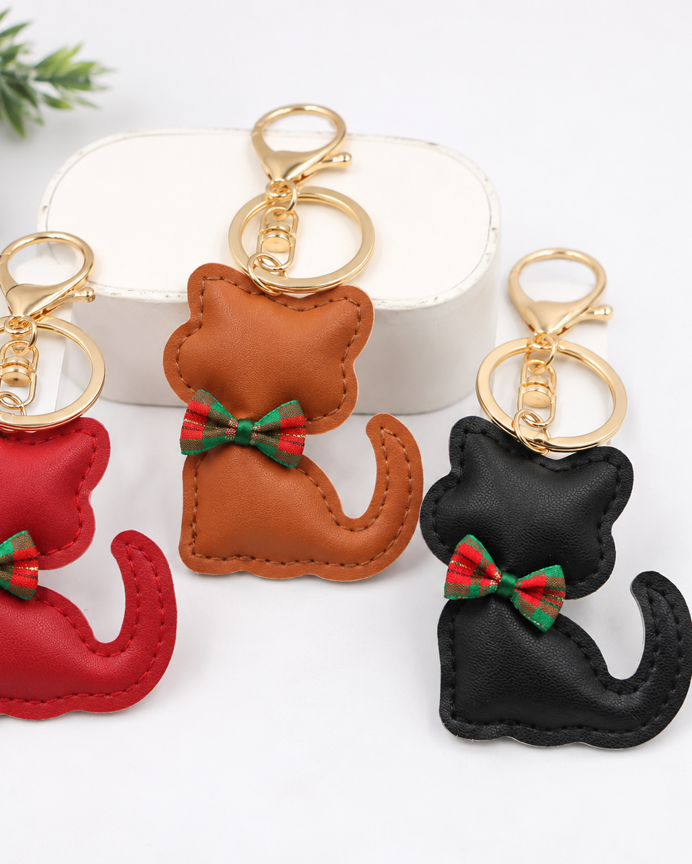 Three faux leather cat-shaped keychains with gold-tone keyrings and red, green, and white bow accents, displayed on a white background with a decorative white box and holiday pine branches.