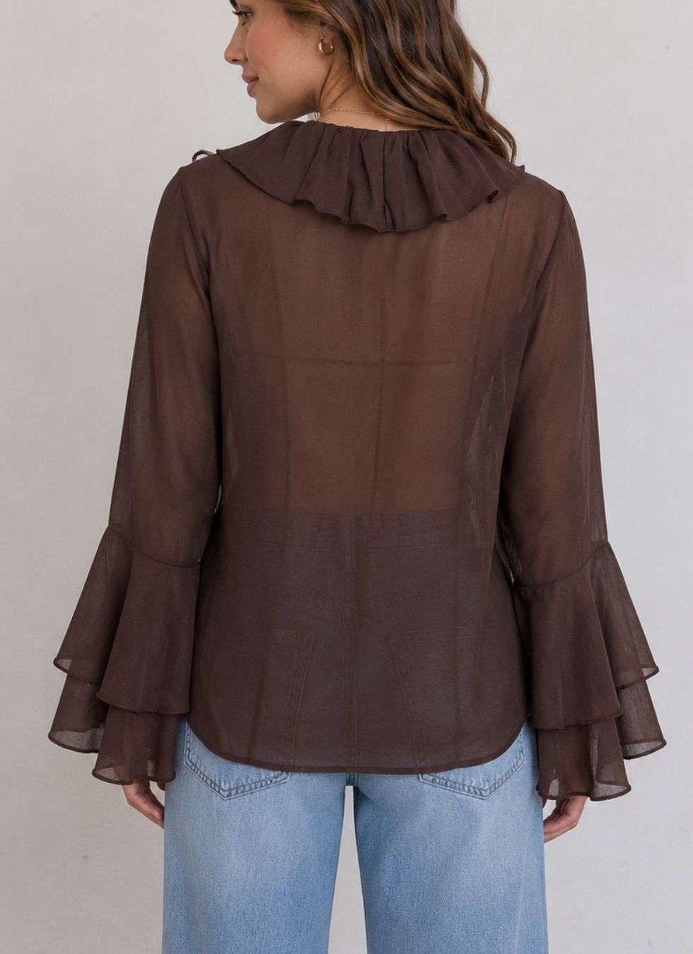 A woman shown from the back wearing a brown sheer long-sleeve blouse with a ruffled collar and layered ruffle cuffs, paired with light blue jeans, against a plain light gray background.