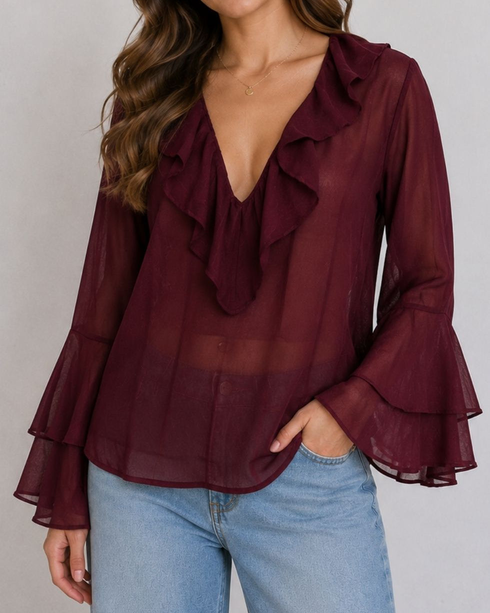 A woman wearing a sheer burgundy long-sleeve blouse with a deep V-neckline, layered ruffles, and flared bell sleeves, paired with light blue jeans against a neutral gray background.