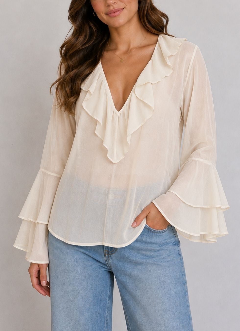 A woman wearing a cream-colored sheer long-sleeve blouse with a ruffled V-neckline and tiered flared sleeves, paired with light blue jeans, against a neutral gray background.