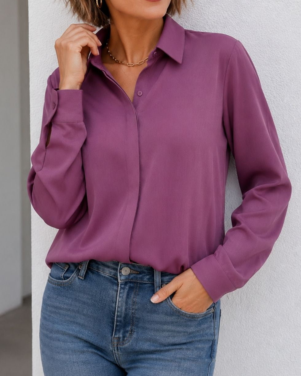 A woman wearing a long-sleeve button-up blouse in a dusty purple/plum color, paired with blue jeans, standing against a light gray textured wall.