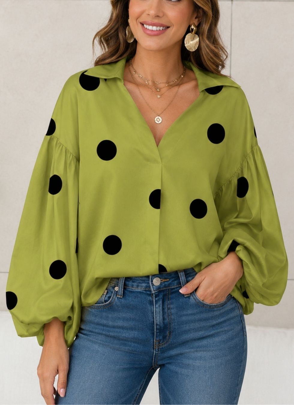 A woman wearing a lime green long-sleeve blouse with black polka dots and blue jeans, shown from the waist up against a light neutral wall.