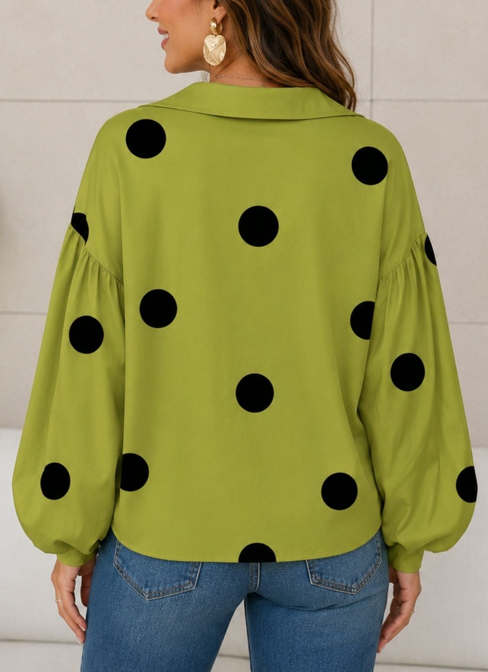A woman wearing a lime green long-sleeve blouse with large black polka dots, shown from the back. She is wearing blue jeans and gold statement earrings against a light gray paneled wall.