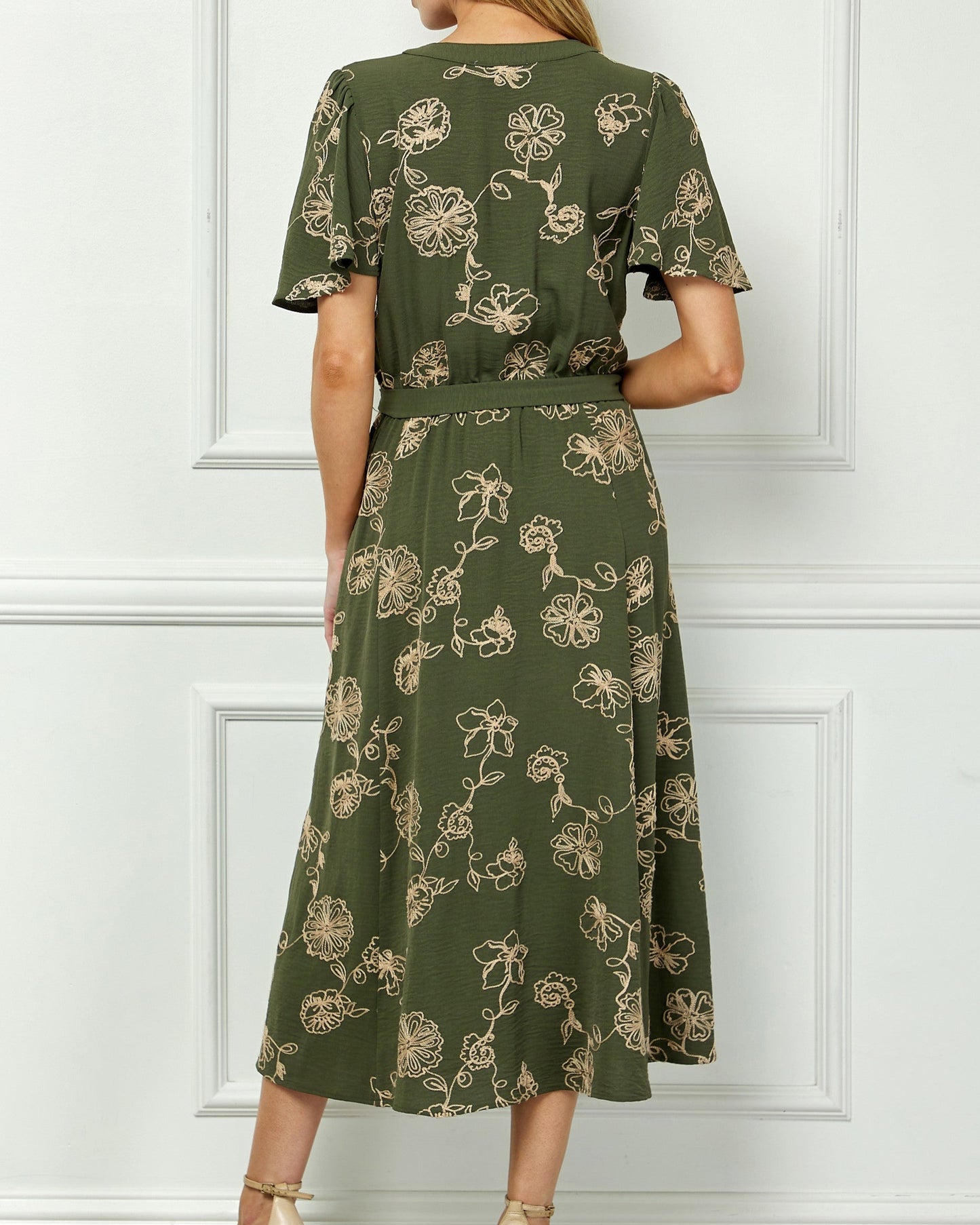 Woman wearing a green floral dress against a white paneled wall.