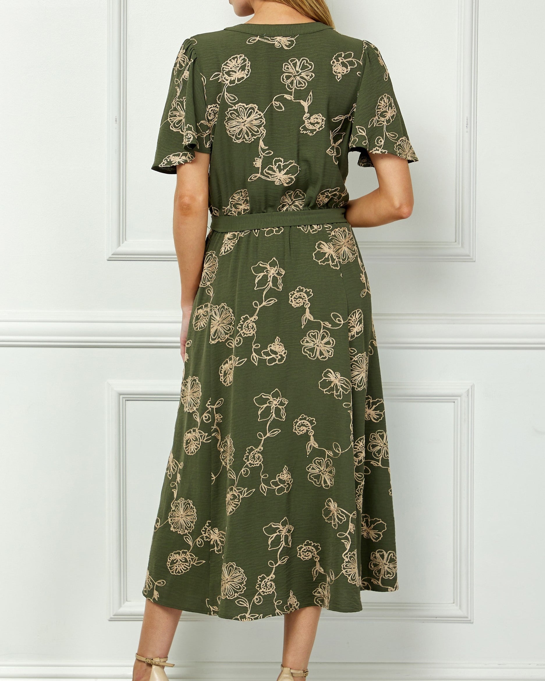 Woman wearing a green floral dress against a white paneled wall.