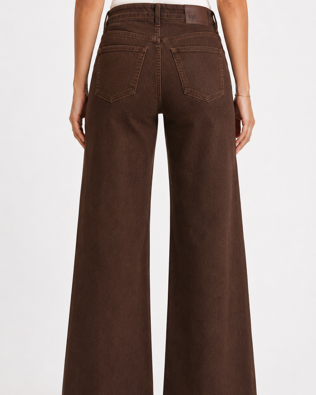A person shown from the back wearing dark brown wide-leg jeans with a white top and tan heels, photographed against a light gray studio background.