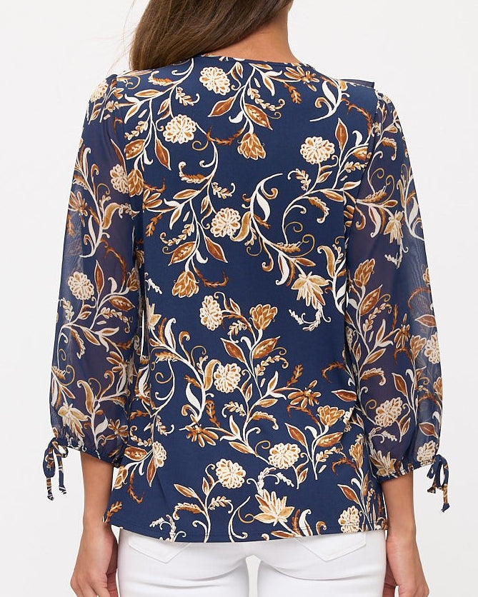 A woman viewed from the back wearing a navy blue blouse with a beige and white floral pattern and white pants, set against a plain white background.