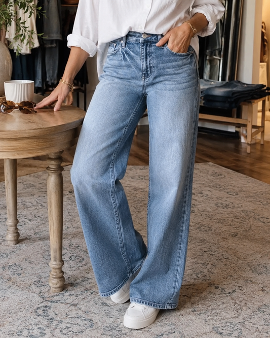 A woman wearing a white button-down shirt and light-wash wide-leg blue jeans stands in a home interior next to a wooden table.