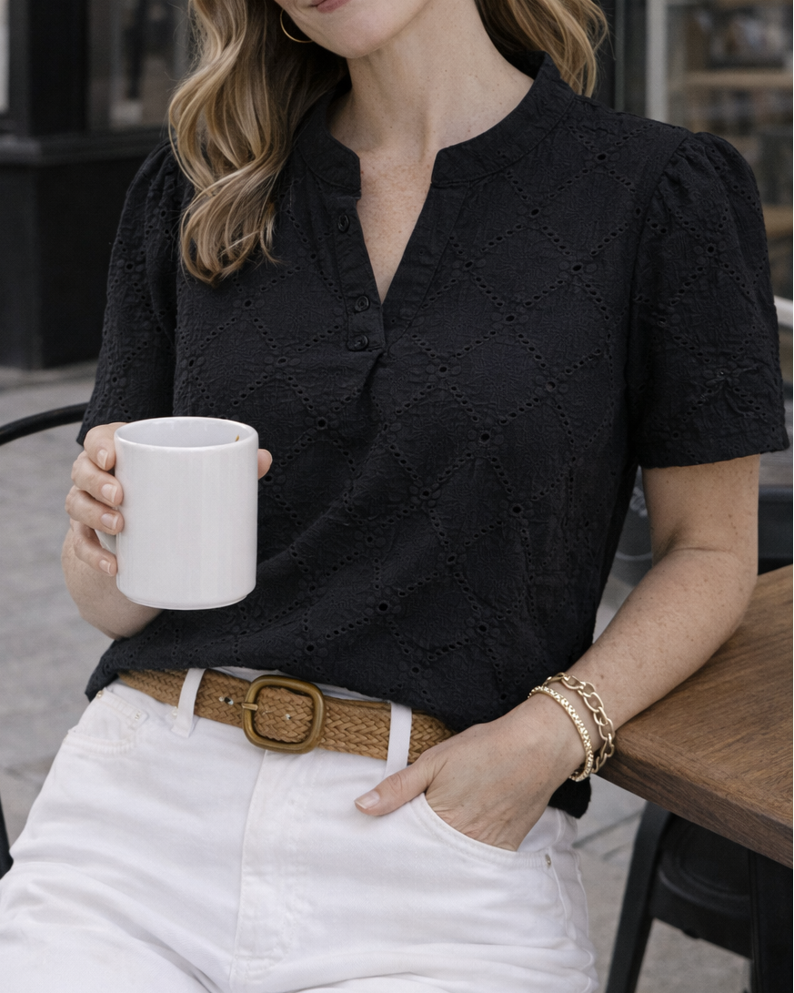 Woman sitting outdoors holding a white mug, wearing a black shirt and white pants.