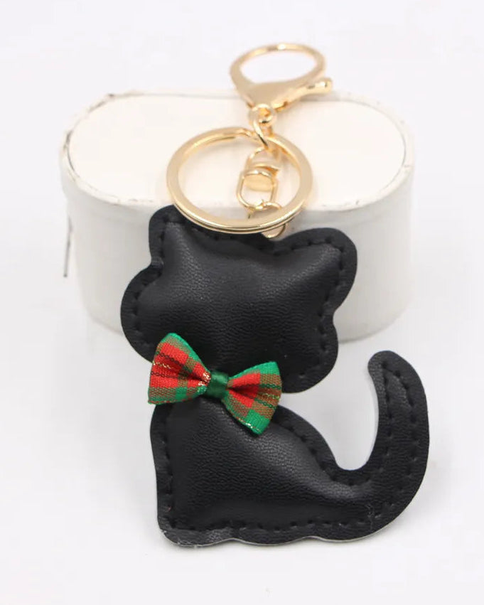 Black cat-shaped keychain bag with a red and green plaid bow tie, attached to gold-tone key rings, shown in front of a white cylindrical display block on a white background.