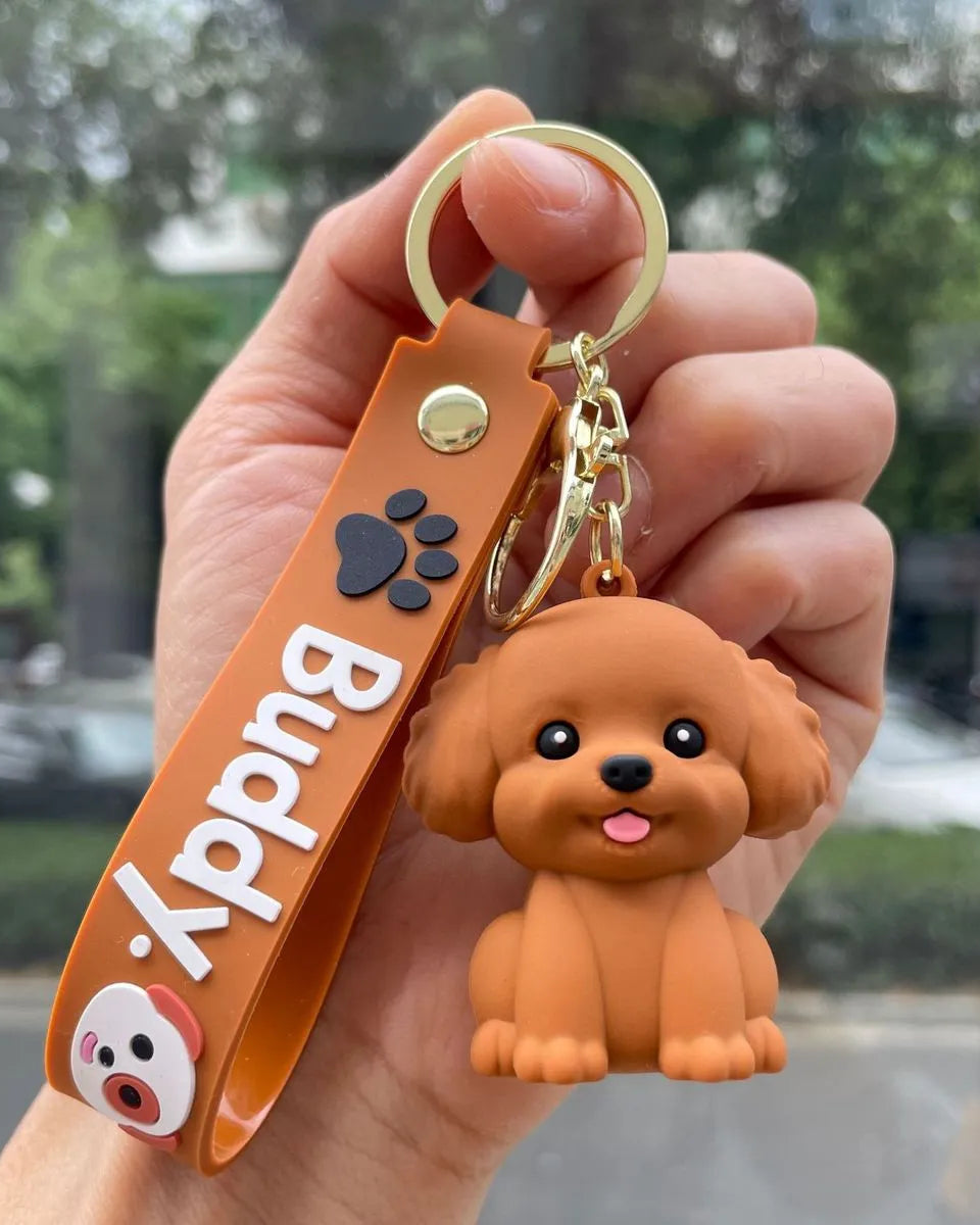 A hand holds a brown rubber keychain featuring a small 3D dog charm and a matching brown strap with the name 'Buddy' in white text, set against a blurred outdoor background.