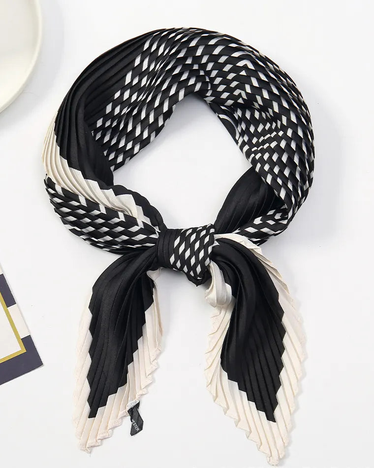A pleated silk scarf with a black and white geometric pattern, tied in a knot and displayed on a clean white surface with a small decorative card and plate in the background.