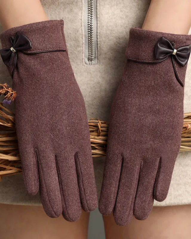 A pair of women's knit gloves in a muted mauve-brown color, featuring a black faux-leather bow with a small silver rhinestone accent at the wrist. The gloves are shown on a model wearing a beige coat, with dried flowers resting on the coat.