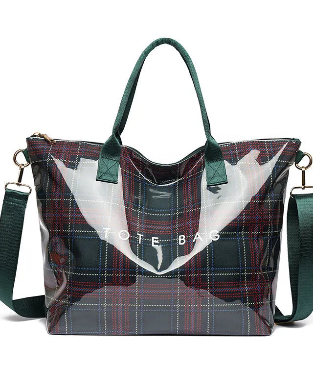 A tote bag with a transparent vinyl front, a red and blue plaid patterned body, dark green handles and shoulder strap, and gold-tone hardware, shown on a white background.