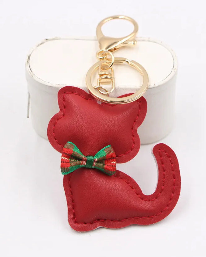 A red faux-leather cat-shaped keychain with a green and red striped bow, attached to gold-tone rings, displayed in front of a white cylindrical prop on a white background.