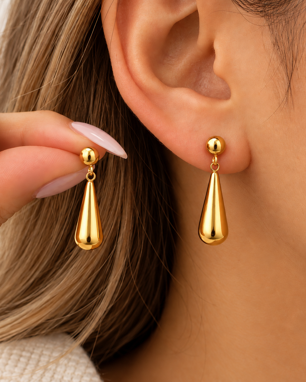 A close-up of a person wearing shiny gold teardrop-shaped drop earrings, with one earring on the earlobe and the other held near the ear against a soft light background.