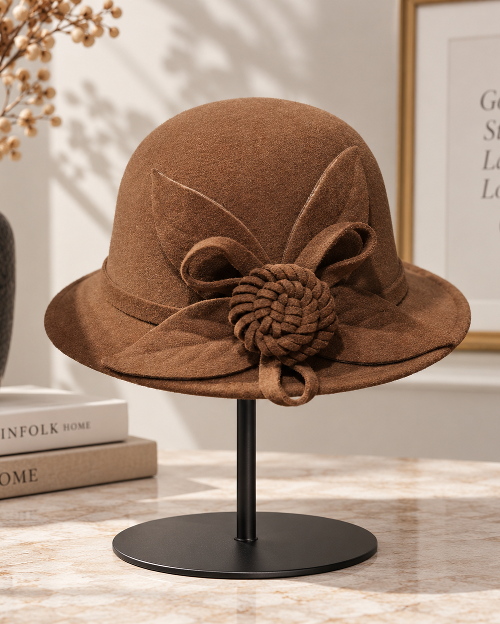 A brown felt cloche hat with a decorative bow and knotted pom-pom accent on the brim, displayed on a black stand on a marble tabletop with a vase, books, and framed wall art in the background.
