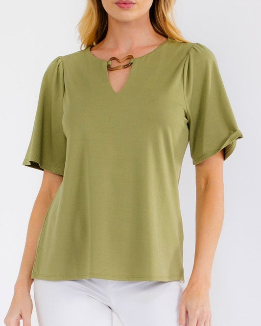 A woman modeling a short-sleeve olive green top with a keyhole cutout at the neckline, paired with white cropped pants and clear high-heeled sandals, against a plain white studio background.